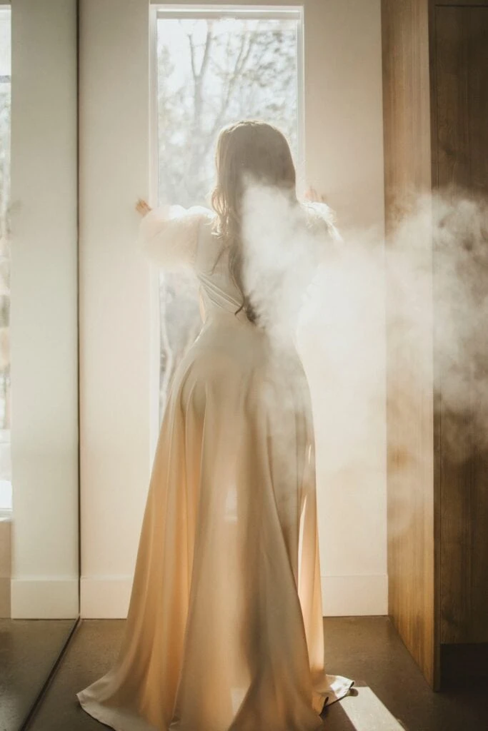 Ava Marie, clad in an exquisite dress, stands in a doorway, her back to the viewer. Sunlight pours about her form and an enigmatic fog billows behind her, sketching a fantastical and celestial ambiance. Ava Marie Halifax's Elite Independent Companion